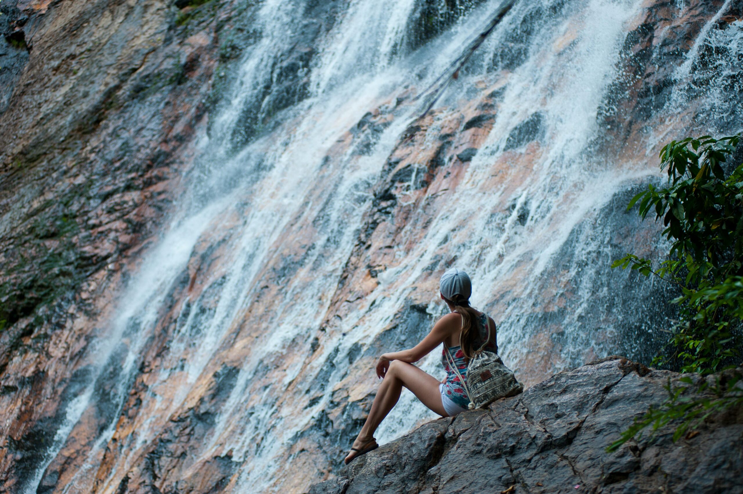 A woman sits on rocks by a cascading waterfall in Thailand, embracing adventure and nature.